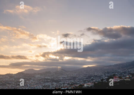 Den Blick über die weitläufigen Hügel Stadt Funchal mit Bergen in der Ferne. Sonnenuntergang mit den letzten Strahlen der Sonne durch die Wolken brechen Stockfoto