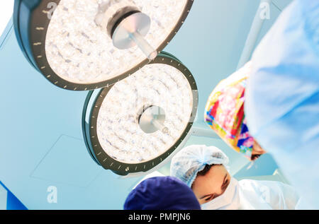 Eine weibliche Chirurgen steht unter einem chirurgischen Lampe in Betrieb Zimmer während der Durchführung einer Operation auf einem blauen Hintergrund Stockfoto