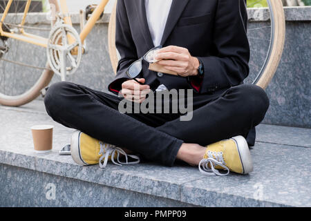 Stilvolle Mann im schwarzen Anzug und Freizeitschuhe seine Lesung Brillen im freien Reinigung. Hipster sitzende Person im städtischen Bereich mit Tasse Kaffee vor Stockfoto