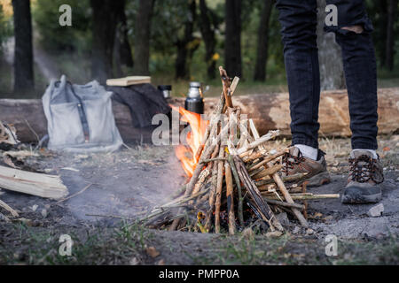 Brennen Lagerfeuer in der Nähe von einem Wald. Reisen in das wilde Konzept: Campingplatz mit Vintage Rucksack, Thermoskannen und männliche Füße in Trekking Schuhe in der Nähe von eine Stockfoto
