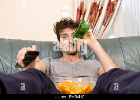 Junger Mann mit der Fernbedienung und Bier und Chips auf der Couch aufgeregt Stockfoto