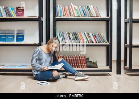 Portrait der Junge gut aussehende Schüler Mädchen mit kurzen blonden Haaren in lässig-elegante Kleidung sitzen auf Fuss in der modernen Bibliothek in der Nähe von Regalen, lesen Lieblingsbücher, Ausgaben Wochenende in gemütlicher Atmosphäre. Stockfoto