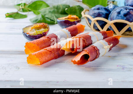 Frisches Lila Pflaumen in einem korbwaren Vase und auf dem Tisch verstreut, Pflaumen in Form von trockenen Späne und Obst Leder rollt auf weißen Holz- Hintergrund. D Stockfoto