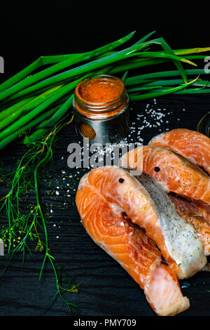 Scheiben der rote Fisch Lachs auf Forelle mit frischen Frühlingszwiebeln, Petersilie, peppper, Gewürze, Dill, rosa und weißen Salz, selery Grüns auf schwarzen Holz- backgrou Stockfoto