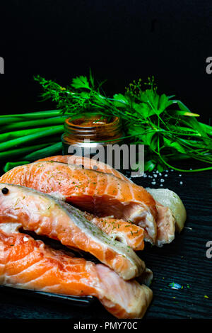 Scheiben der rote Fisch Lachs auf Forelle mit frischen Frühlingszwiebeln, Petersilie, peppper, Gewürze, Dill, rosa und weißen Salz, selery Grüns auf schwarzen Holz- backgrou Stockfoto