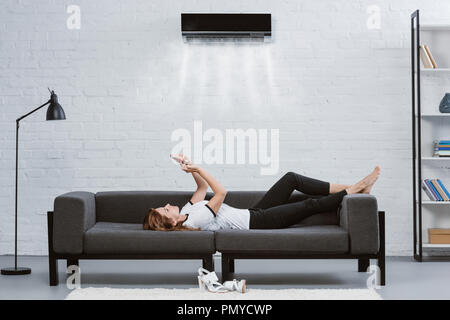 Entspannt junge Frau mit Smartphone auf der Couch unter der Klimaanlage an der Wand hängend Stockfoto