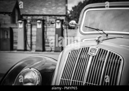 Schwarz-Weiß, Nahaufnahme des klassischen Oldtimers Morris Eight (Frontgrill, Scheinwerfer), Vintage-Benzinpumpen im Hintergrund. Stockfoto