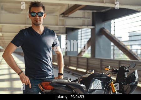 Portrait von attraktiven Biker neben Motorrad am Parkplatz. Im städtischen Hintergrund. Stockfoto