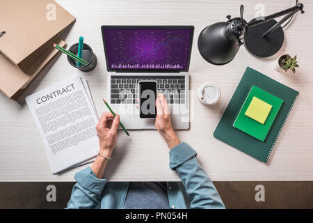 Zugeschnittenes Bild der Geschäftsmann mit Smartphone mit Vertrag, Schreibwaren und Laptop mit Grafik auf dem Bildschirm Stockfoto