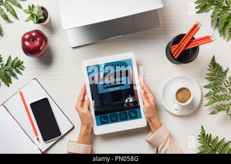 Zugeschnittenes Bild der weiblichen Freiberufler Holding digital Tablet mit Buchung auf dem Bildschirm zu Tisch mit Gadgets und Kaffee von farnblätter umgeben Stockfoto