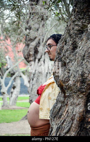 Schwangere Frau ihr Gesicht verstecken hinter einem Baum und Freund aus ihrem Gesicht, lustiges Foto von Schwangeren, Witz Stockfoto