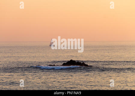 Ein entfernter Segelboot Segeln entlang dem Meer bei Sonnenuntergang mit einigen Wellen auf einem Felsen, mit dem orangefarbenen Himmel im Hintergrund. Stockfoto