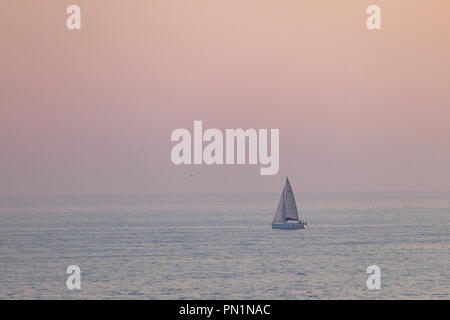 Ein entfernter Segelboot Segeln entlang dem Meer bei Sonnenuntergang, mit der rosa gefärbten Himmel im Hintergrund. Stockfoto