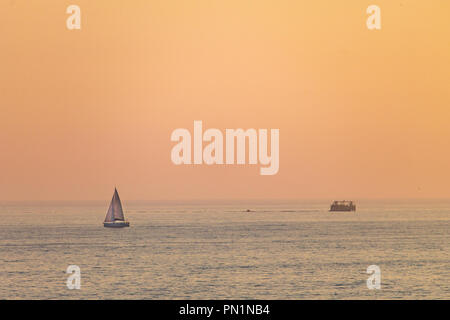 Ein entfernter Segelboot Segeln entlang dem Meer bei Sonnenuntergang, mit dem orangefarbenen Himmel im Hintergrund. Stockfoto