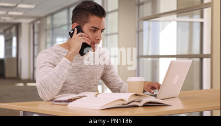 Tausendjährige Latino Student in Campus Bibliothek mit Laptop und Mobiltelefon Stockfoto