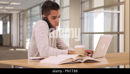 Tausendjährige Latino Student in Campus Bibliothek mit Laptop und Mobiltelefon Stockfoto