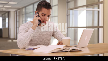 Tausendjährige Latino Student in Campus Bibliothek mit Laptop und Mobiltelefon Stockfoto