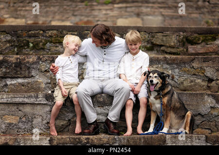 Eine junge, liebevoller Vater ist draußen sitzen auf Steinstufen, mit seinen zwei jungen Kinder und ihre angenommene Haustier Deutscher Schäferhund an einem Tag im Sommer. Stockfoto