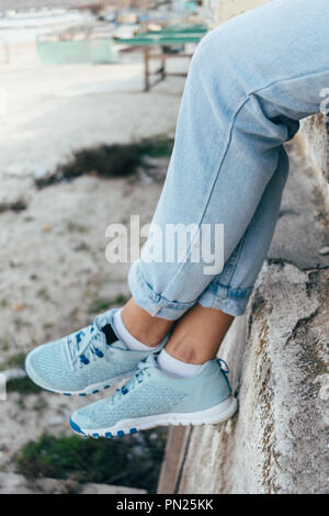 In der Nähe der weiblichen Beine in Blau, Jeans und Turnschuhe. Junge Frau sitzt auf hohe Beton Brüstung in der Stadt, vertikale Framing. Stockfoto