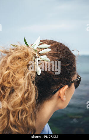 Close-up Curly blonde junge Frau mit wenig Stammzellen in ihrem Pferdeschwanz sitzen auf dem Meer. Vertikale Foto unkenntlich Weibliche trägt blaue Shirt Stockfoto