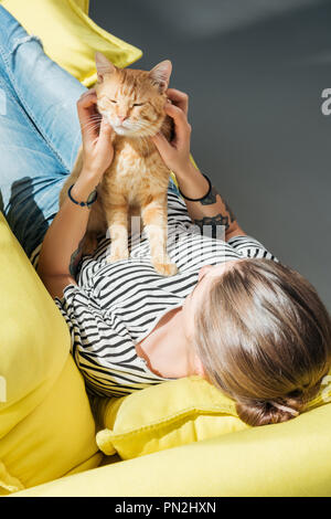 Hohe Betrachtungswinkel von Mädchen spielen mit niedlichen rote Katze beim Lügen auf Gelb couch Stockfoto