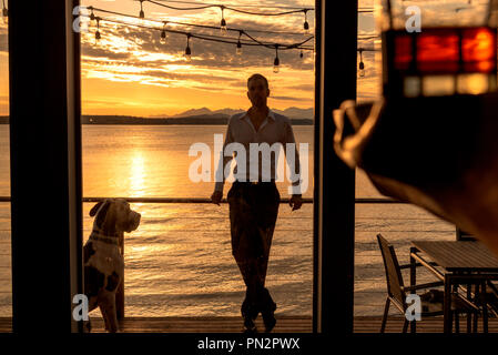 Silhouetted Mann mit Dogge Hund auf Deck in Beach home mit einem Glas Alkohol im Vordergrund bei Sonnenuntergang. Stockfoto