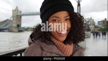 Gerne schwarze Frau lächelnd vor der Tower Bridge tragen Jacke und Mütze Stockfoto