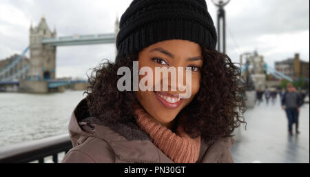 Gerne schwarze Frau lächelnd vor der Tower Bridge tragen Jacke und Mütze Stockfoto