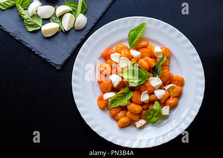 Gnocchi alla Sorrentina in Tomatensauce mit frischem Basilikum und Mozzarella Kugeln auf ein weißes Schild mit schwarzem Schiefer Schneidbrett serviert. Tra Stockfoto