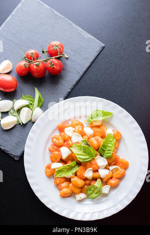 Gnocchi alla Sorrentina in Tomatensauce mit frischem Basilikum und Mozzarella Kugeln auf Teller serviert und Zutaten auf schiefer Board auf Schwarz b Stockfoto