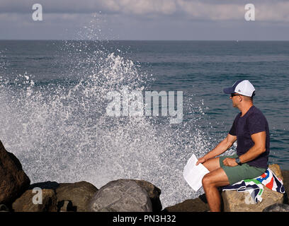 Ein Mann sitzt am Rande beobachten Wellen des Ozeans an den Felsen neben ihm aus dem Pazifischen Ozean im Süden von Kalifornien, USA Stockfoto