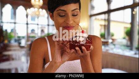Schwarze Frau nimmt chaotisch Bissen von Red velvet Cupcake im Innenbereich Stockfoto