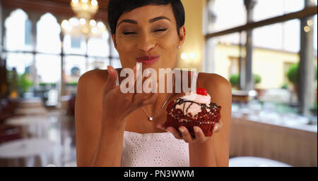 Schwarze Frau nimmt chaotisch Bissen von Red velvet Cupcake im Innenbereich Stockfoto