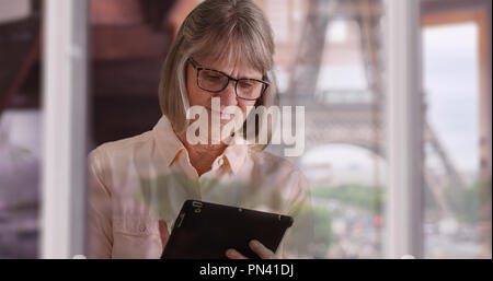 Reife Lady mit Pad Gerät im Innenbereich mit Eiffelturm Reflexion über Fenster Stockfoto