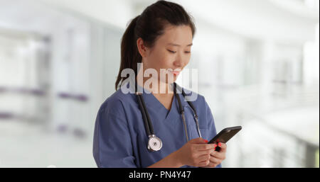 Glückliche junge asiatische Arzt in Scrubs texting mit Smartphone im inneren Leere Krankenhaus Stockfoto