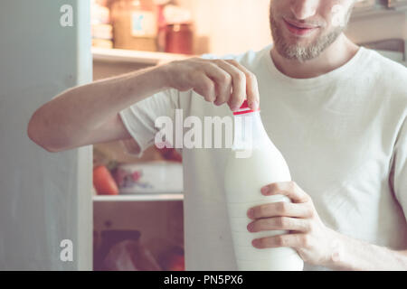 Junger bärtiger Mann öffnet die Flasche Milch stehen in der Nähe der Kühlschrank zu Hause Stockfoto