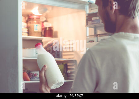 Junger bärtiger Mann öffnet die Flasche Milch stehen in der Nähe der Kühlschrank zu Hause Stockfoto