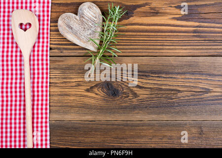 Ein Löffel aus Holz mit einem Ausschnitt in Herzform, Zweige Rosmarin und ein Herz aus Holz liegen auf einer rot-weiß karierten Tuch auf einem rustikalen Holzmöbeln Hintergrund Stockfoto