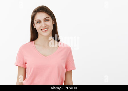 Studio shot der gut aussehende freundlich und unbeschwerte junge Mädchen mit braunem Haar in ungezwungener Rosa t-shirt, freudig lächelnd und Neigekopf mit Interesse, sich entspannt und über graue Wand optimistisch Stockfoto