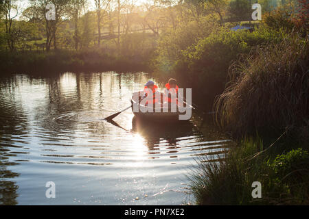 Zwei Jungen rudern ein Boot auf einem kleinen See zurück von der Abendsonne beleuchtet. Stockfoto