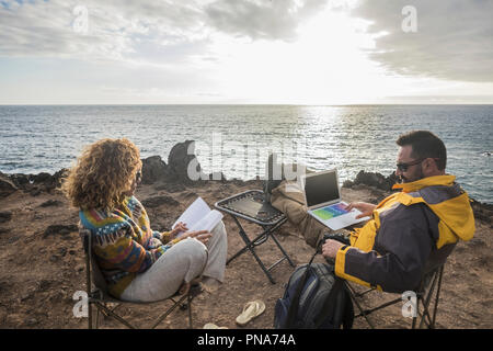 Paar alternativen Reisenden sitzen auf den Felsen am Meer und den Sonnenuntergang genießen die Freiheit und die Arbeit mit einem Laptop oder beim Lesen eines b Stockfoto