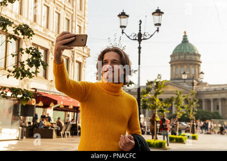 Der junge Mann jemand warten und selfie, er kleidete sich in einen gelben Pullover, einen schwarzen Regenmantel oder Jacke, Jeans, Street und Kazanskiy Kathedrale Stockfoto