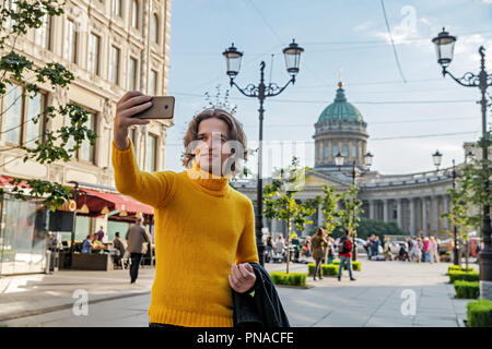 Der junge Mann jemand warten und selfie, er kleidete sich in einen gelben Pullover, einen schwarzen Regenmantel oder Jacke, Jeans, Street und Kazanskiy Kathedrale Stockfoto