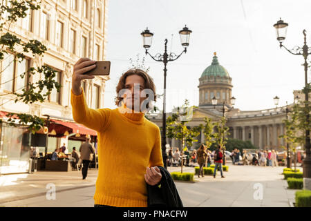 Der junge Mann jemand warten und selfie, er kleidete sich in einen gelben Pullover, einen schwarzen Regenmantel oder Jacke, Jeans, Street und Kazanskiy Kathedrale Stockfoto