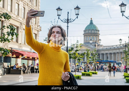 Der junge Mann jemand warten und selfie, er kleidete sich in einen gelben Pullover, einen schwarzen Regenmantel oder Jacke, Jeans, Street und Kazanskiy Kathedrale Stockfoto