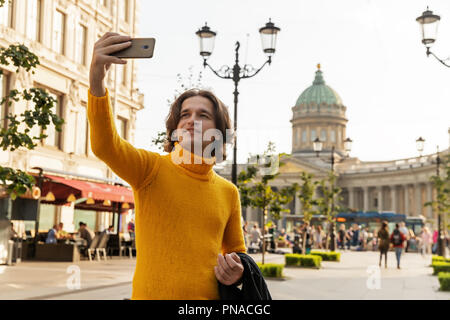 Der junge Mann jemand warten und selfie, er kleidete sich in einen gelben Pullover, einen schwarzen Regenmantel oder Jacke, Jeans, Street und Kazanskiy Kathedrale Stockfoto