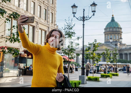 Der junge Mann jemand warten und selfie, er kleidete sich in einen gelben Pullover, einen schwarzen Regenmantel oder Jacke, Jeans, Street und Kazanskiy Kathedrale Stockfoto