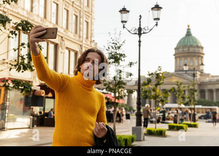 Der junge Mann jemand warten und selfie, er kleidete sich in einen gelben Pullover, einen schwarzen Regenmantel oder Jacke, Jeans, Street und Kazanskiy Kathedrale Stockfoto