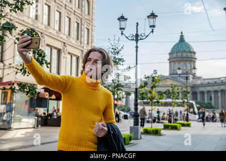 Der junge Mann jemand warten und selfie, er kleidete sich in einen gelben Pullover, einen schwarzen Regenmantel oder Jacke, Jeans, Street und Kazanskiy Kathedrale Stockfoto