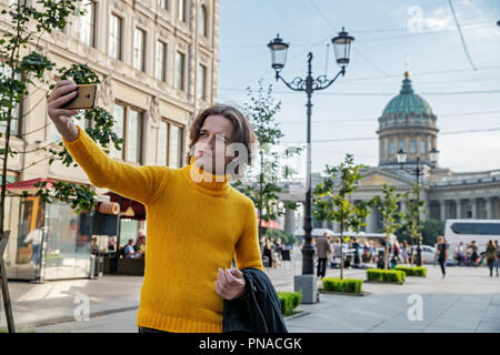 Der junge Mann jemand warten und selfie, er kleidete sich in einen gelben Pullover, einen schwarzen Regenmantel oder Jacke, Jeans, Street und Kazanskiy Kathedrale Stockfoto